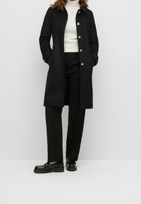 Cappotto nero con colletto, bottoni frontali e tasche laterali, abbinato a pantaloni neri e scarpe nere lucide.