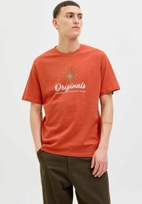 Rust-orange bomulds t-shirt med et hvidt script "Originals" logo og en gylden stjernespænde grafik, afslappet pasform, korte ærmer.