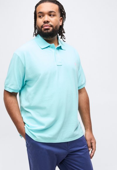 Άνδρας με γενειάδα και κοτσιδάκια, φοράει ανοιχτό μπλε πόλο πουκάμισο και σκούρο μπλε παντελόνι, κοιτάζει προς τα πλάγια με το ένα χέρι στην τσέπη.