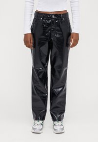 Modell som bär blanka svarta fusk läderbyxor med krokodiltextur, ihop med vita sneakers med gröna accenter och en vit långärmad topp.