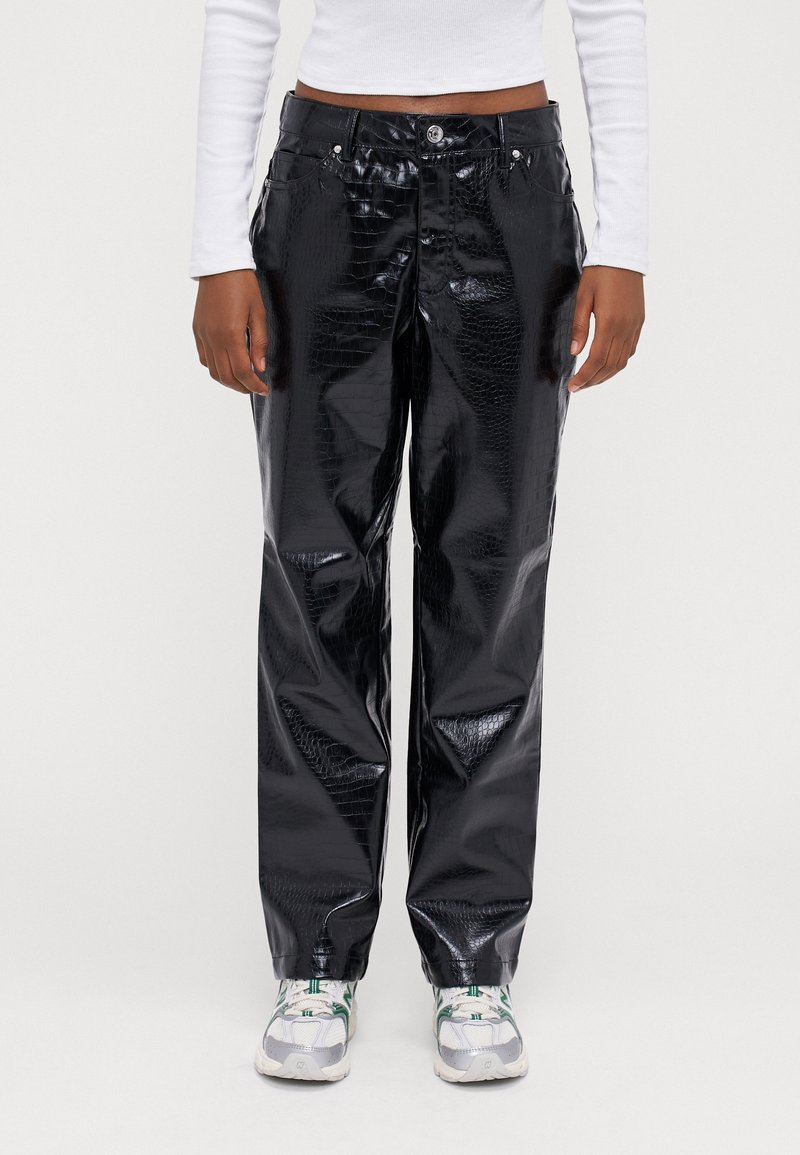 Modell som bär blanka svarta fusk läderbyxor med krokodiltextur, ihop med vita sneakers med gröna accenter och en vit långärmad topp.