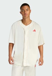 Maglia da baseball bianca in rete con maniche corte, scollo a V, dettagli rossi e logo. Presenta una chiusura a bottone e spacchi laterali per comfort e vestibilità.