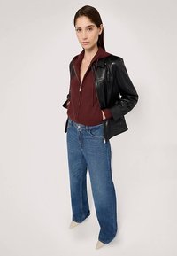 Svart läderjacka över en vinröd zip-up hoodie, kombinerad med ljusblå jeans med raka ben och beige spetsiga klackskor, på en neutral bakgrund.