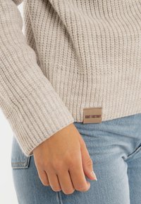 Beige gebreide trui met ribbelstructuur, voorzien van een geweven label aan de zoom. Gecombineerd met lichtblauwe denim jeans.