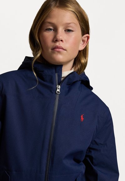 Marineblaue Zip-Jacke mit Kapuze und einem roten gestickten Logo. Sie besteht aus einem Stoff mit glatter Textur und verfügt über zwei Taschen auf Hüfthöhe.