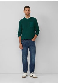Groene gebreide trui met een witte kraag en zak, gecombineerd met blauwe jeans en witte sneakers. Effen achtergrond, frontaanzicht.