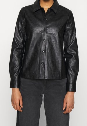 Persona que lleva una camisa abotonada de textura de cuero sintético con diseño de cocodrilo en negro, con mangas largas y pantalones oscuros.