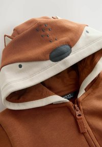 Rust-colored zip-up hoodie featuring a cream hood with fox face design, embroidered nose, and ear accents. Soft fabric texture.
