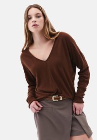 Brauner V-Ausschnitt-Pullover aus weichem Material, mit langen Ärmeln und gerippten Bündchen, kombiniert mit einem grauen Rock und einem goldenen Gürtel mit Kettendetails.