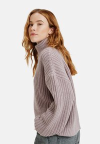 Pull en maille épaisse de couleur violet clair avec un col montant, des manches larges et une texture côtelée. Le design présente une coupe décontractée et des fentes sur les côtés.