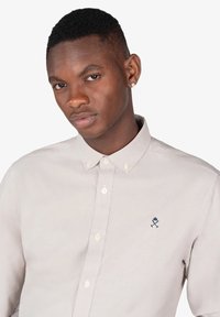 Camisa de botones gris claro con cuello clásico, mangas cortas y un acento bordado en azul marino en el pecho. Textura de tela suave.