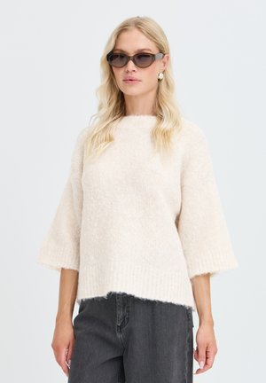 Femme portant des lunettes de soleil foncées, des boucles d'oreilles argentées, un pull crème duveteux à manches larges, et un jean gris foncé sur un fond uni.