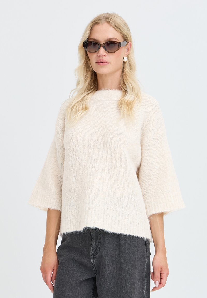 Femme portant des lunettes de soleil foncées, des boucles d'oreilles argentées, un pull crème duveteux à manches larges, et un jean gris foncé sur un fond uni.