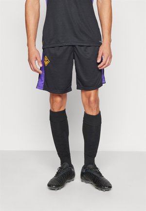 Athlète masculin portant un short de football noir et violet, des chaussettes noires montantes jusqu'aux genoux, et des crampons noirs, debout devant un fond uni.