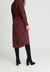 Abito lungo a zigzag rosso e nero con maniche lunghe, vita cinturata, abbinato a stivali neri con tacco alti sopra il ginocchio.