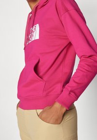 Persona che indossa un felpa rosa brillante con un logo bianco, mani nelle tasche dei pantaloni beige, sezione centrale accorciata con uno sfondo neutro.
