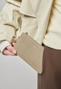 Beige Lederportemonnaie mit mehreren Kartenfächern, Reißverschluss und geprägtem Markennamen. Glatte Oberfläche, rechteckige Form.