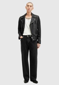 Schwarze Leder-Motorradjacke mit Nietenakzenten, weißes T-Shirt, lockere schwarze Jeans und schwarze Schuhe; stehende Pose vor neutralem Hintergrund.