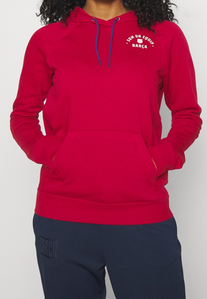 Červená mikina s kapucí, s kapsou typu klokan, modrými šňůrkami a nápisy „SOM UN EQUIP“ a „BARÇA“ potištěnými bílou barvou na hrudi. Materiál je měkký.