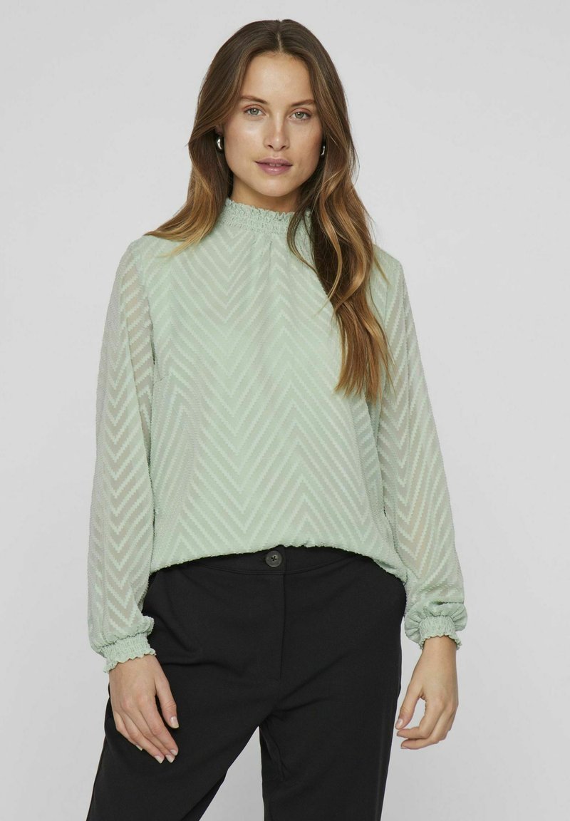Mint green long-sleeve blouse with a textured chevron pattern, elasticized cuffs, and a gathered neckline, paired with black pants.