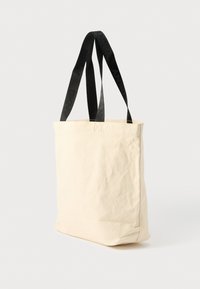 Béžová plátená taška s čiernymi nylonovými popruhmi. Popruhy majú logo v opakujúcom sa texte. Pevný dizajn s obdĺžnikovým tvarom a jemnou textúrou.