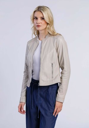 Veste bombardier en cuir beige clair avec un col rond, deux poches zippées à l'avant et une texture lisse, associée à un haut blanc ajusté et un pantalon bleu.