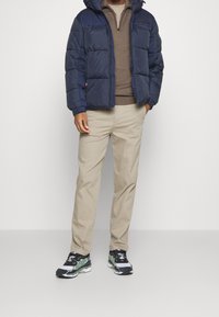 Marinblå puffjacka med huva, beige tröja, ljusbeiga byxor och svarta/gröna sneakers. Har quiltad design och dragkedjestängning.
