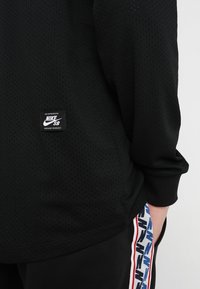 Svart långärmad topp med perforeringar och en vävd Nike SB-etikett. Har ribbade muddar och sidopanel med ett kontrasterande logorandigt mönster.