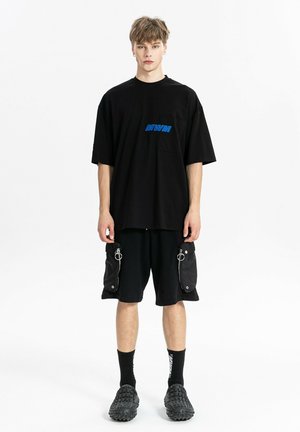 Joven con camiseta negra extragrande con texto azul, pantalones cortos cargo negros con bolsillos con cremallera, calcetines negros y zapatos negros texturizados, de pie.