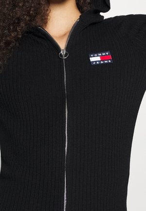 Gros plan d'une personne portant un pull zippé côtelé noir avec un écusson logo Tommy Jeans sur la poitrine gauche.