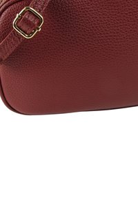 Bordeauxroter Ledertasche mit strukturierter Oberfläche, die eine goldene Schnalle als Akzent am Riemen hat. Runde Basis und glatte Kanten.