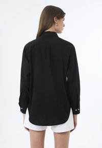 Schwarzes Langarmshirt mit geradem Schnitt, einer einzelnen Rückenfalte und geknöpften Manschetten. Über weißen Shorts getragen, mit glatter Stoffoberfläche.