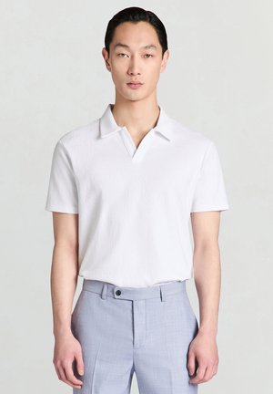 Uomo che indossa una polo bianca a maniche corte dalla trama strutturata, infilata in pantaloni su misura grigio chiaro, in piedi davanti a uno sfondo neutro.