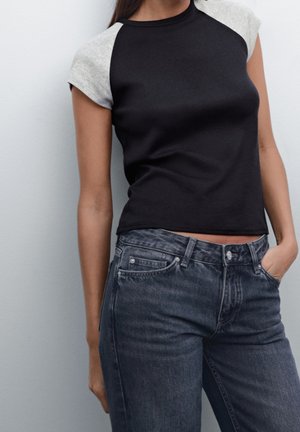 Femme portant un haut noir à manches courtes avec des manches grises et un jean noir délavé taille haute, posant avec une main dans la poche.