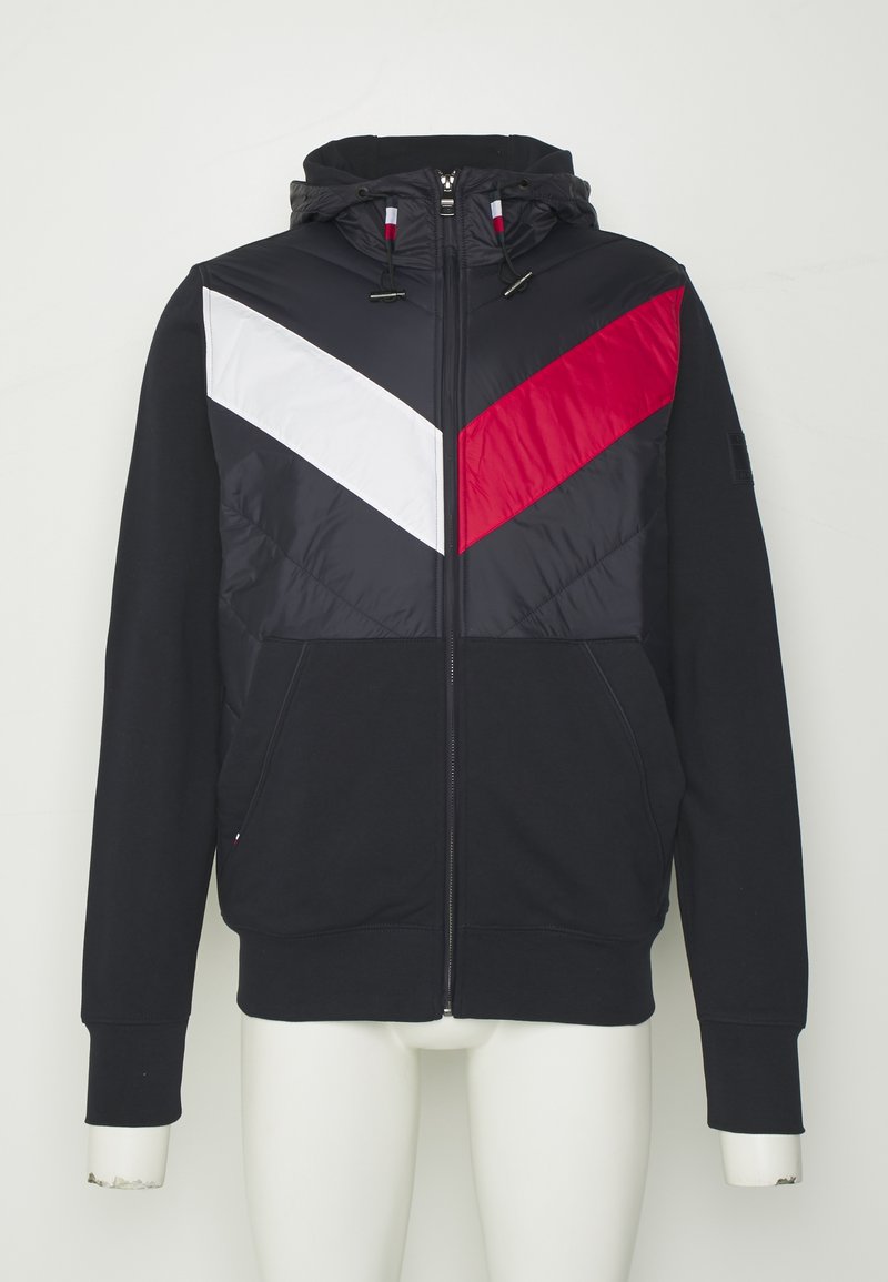 Felpa con cappuccio e chiusura a zip, realizzata in tessuto blu navy, con una parte frontale imbottita decorata da strisce diagonali bianche e rosse, e tasche laterali.