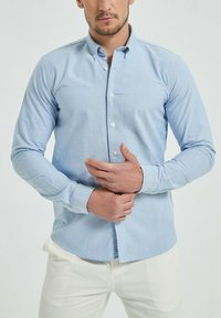 Chemise à manches longues bleu clair avec un col boutonné, fabriquée dans un tissu doux. Présente une fermeture à boutons sur le devant et un ourlet droit.