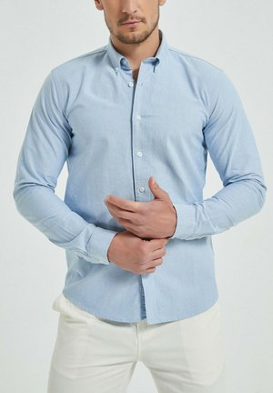 Chemise à manches longues bleu clair avec un col boutonné, fabriquée dans un tissu doux. Présente une fermeture à boutons sur le devant et un ourlet droit.