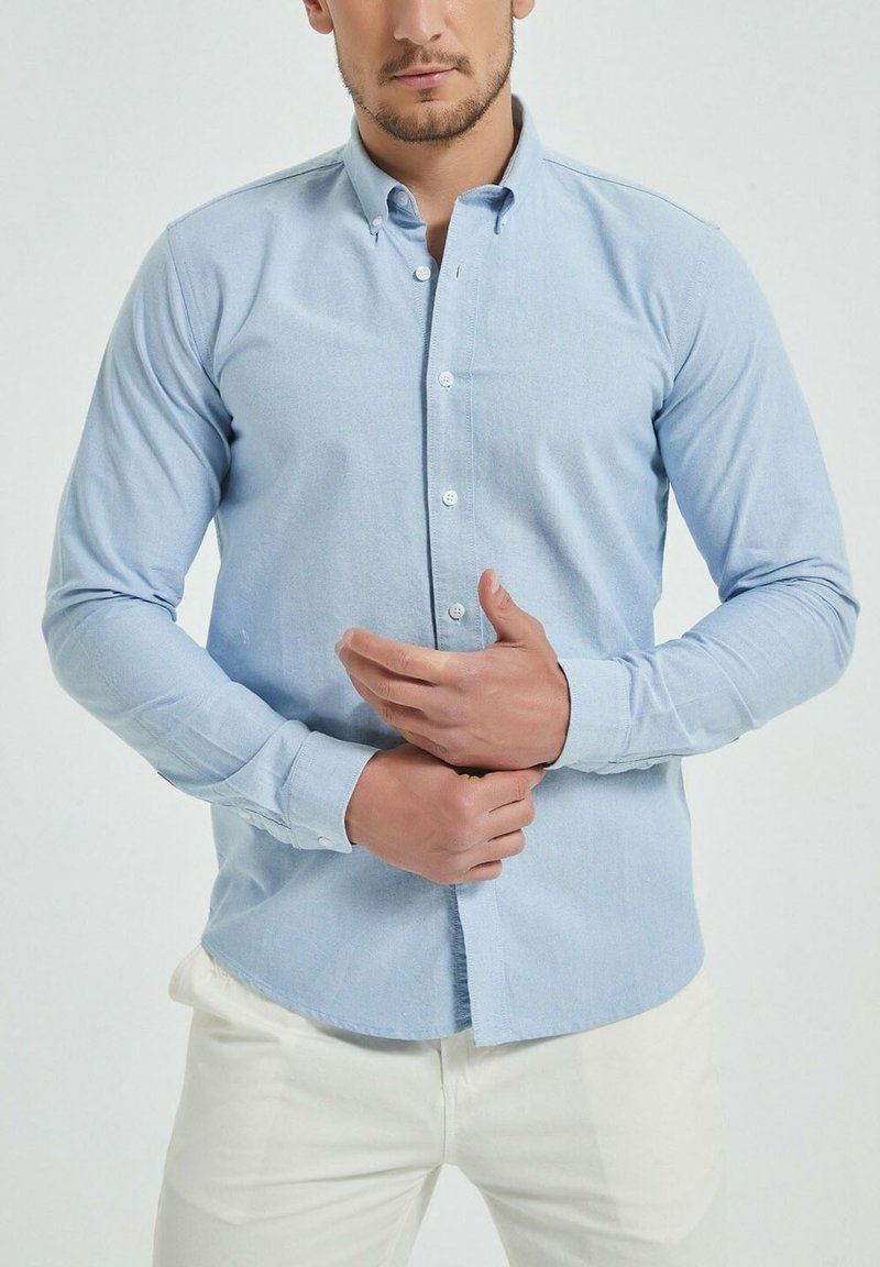 Chemise à manches longues bleu clair avec un col boutonné, fabriquée dans un tissu doux. Présente une fermeture à boutons sur le devant et un ourlet droit.