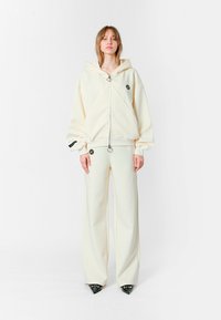 Sudadera blanca con capucha, cremallera frontal y logotipo, combinada con pantalones de pierna ancha a juego. El material parece suave, con textura mínima.