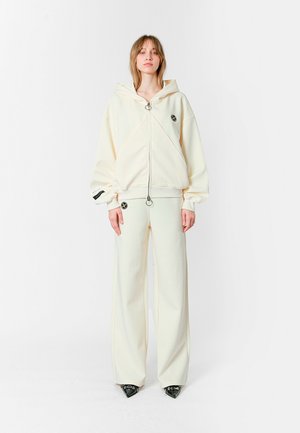 Sudadera blanca con capucha, cremallera frontal y logotipo, combinada con pantalones de pierna ancha a juego. El material parece suave, con textura mínima.