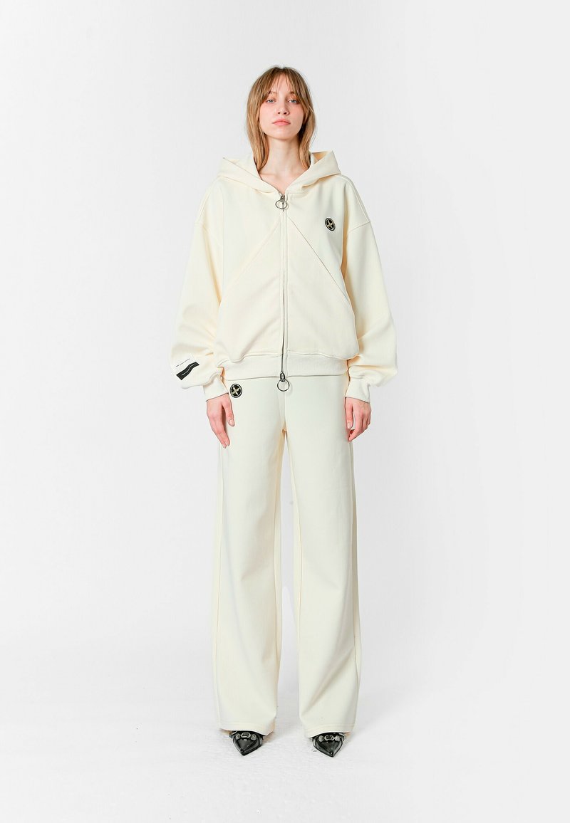 Sudadera blanca con capucha, cremallera frontal y logotipo, combinada con pantalones de pierna ancha a juego. El material parece suave, con textura mínima.