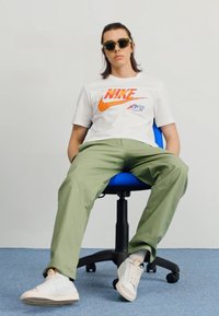 Hvid Nike t-shirt med orange logo; olivengrønne bukser; solbriller; siddende på en blå kontorstol mod en lys baggrund.