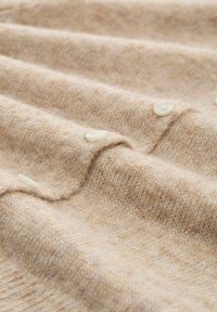 Suéter de punto beige con una textura suave, que presenta varias filas de tela y botones blancos como detalles destacados.