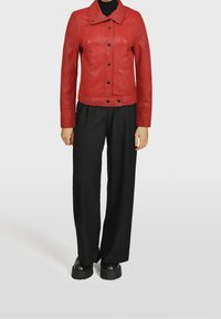 Červená kožená bunda s límcem, předními knoflíky a švy, v kombinaci s volnými černými kalhotami a robustními černými botami.