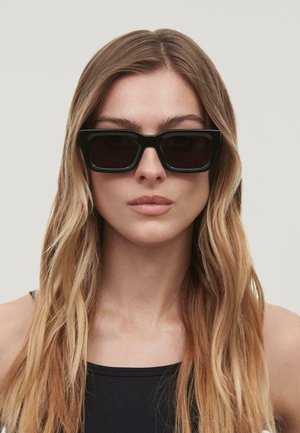 Black rectangular sunglasses with dark lenses, thick frames, and a flat top. Model has long, straight hair with a neutral background.