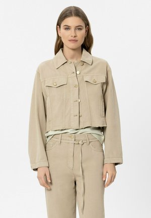 Woman wearing beige cropped jacket with button closures and matching high-waist pants with a tie belt, standing against a plain background.