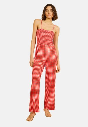 Žena v červeno-bielom kockovanom bezrukávovom overal s širokými nohavicami a tenkými ramienkami stojí s neutrálnym výrazom tváre.