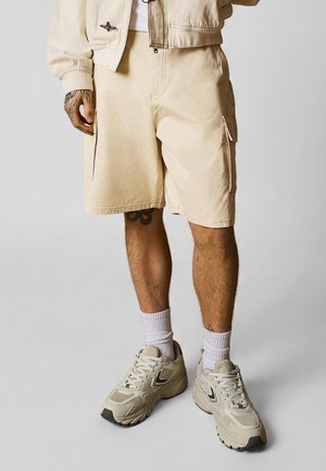 Osoba ubrana w beżowe szorty typu cargo, pasującą kurtkę, białe skarpetki i beżowe masywne sneakersy, stojąca na jednolitym tle.