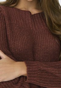 Maglione lavorato a maglia marrone con una texture a costine, collo ampio e maniche lunghe con polsini a costine. Colore ricco e caldo con sottili variazioni.