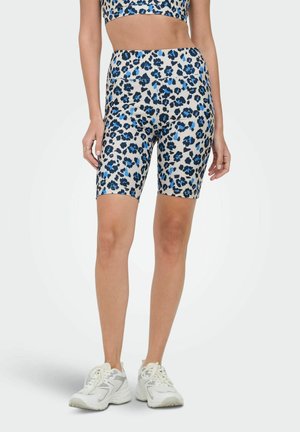 Kobieta w beżowych, wysokowahaczych spodenek rowerowych z niebiesko-czarnym panterkowym nadrukiem oraz białych trampkach, stojąca na jednolitym tle.